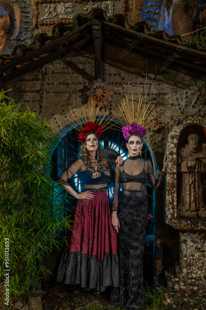Mexican women dressed as catrinas, with a flower headdress on their heads, outside a chapel in a night photo.