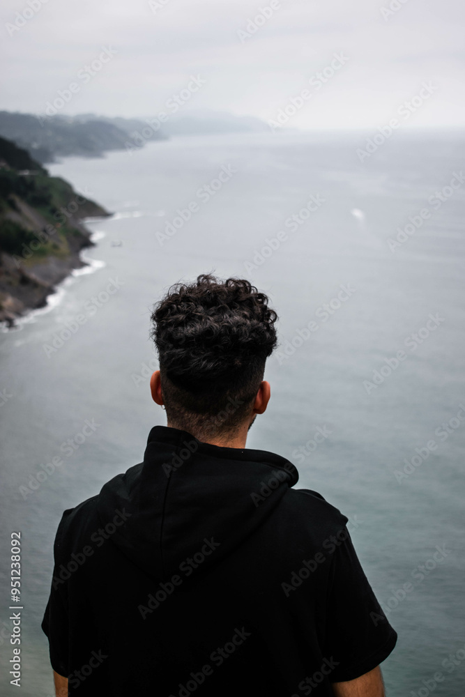 chico joven de pelo rizado negro mirando el mar desde un acantilado en un día de tormenta chico joven de pelo rizado negro mirando el mar desde un acantilado en un día de tormenta
