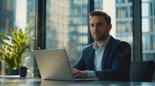 A businessman sits at a sleek desk, deeply engaged with his laptop, surrounded by ambient natural light pouring through large windows, enhancing the modern office vibe.