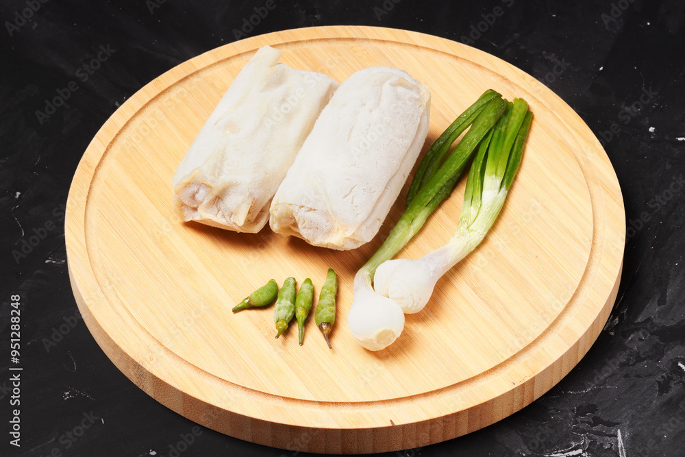 Two frozen lumpia filled with bamboo shoots and meat, with white wrappers, ready to fry. Served with green chili peppers and scallions, photographed on a round wooden tray against a dark background. Two frozen lumpia filled with bamboo shoots and meat, with white wrappers, ready to fry. Served with green chili peppers and scallions, photographed on a round wooden tray against a dark background.