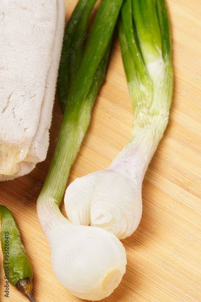 Two frozen lumpia filled with bamboo shoots and meat, with white wrappers, ready to fry. Served with green chili peppers and scallions, photographed on a round wooden tray against a dark background.