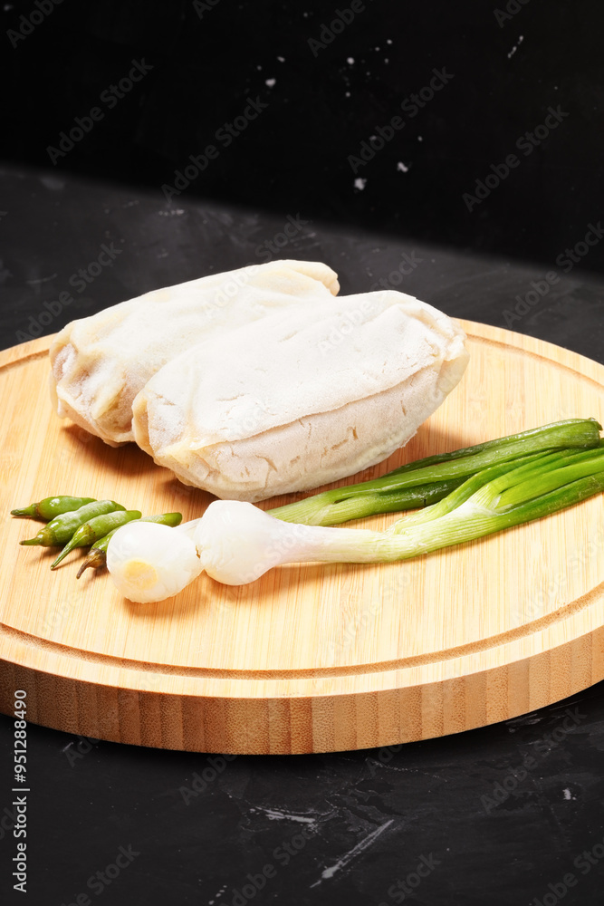 Two frozen lumpia filled with bamboo shoots and meat, with white wrappers, ready to fry. Served with green chili peppers and scallions, photographed on a round wooden tray against a dark background. Two frozen lumpia filled with bamboo shoots and meat, with white wrappers, ready to fry. Served with green chili peppers and scallions, photographed on a round wooden tray against a dark background.