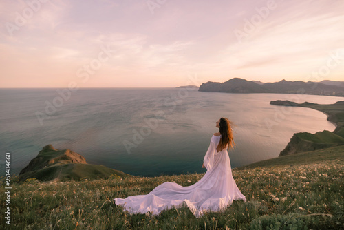 A woman in a white dress stands on a hill overlooking the ocean