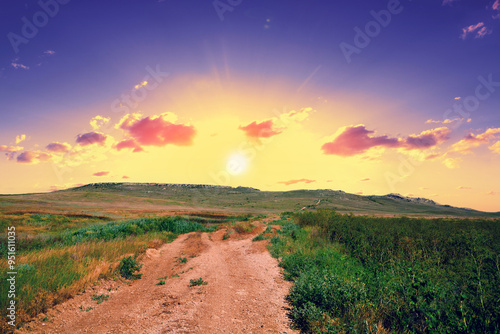 Empty dirt rural road in the meadow to the mountain during sunset photo