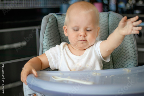 Cute blond caucasian baby child with blue eyes sitting at highchair after eating food. Child is dirty and everything is in rice grains. Baby is serious and surprised