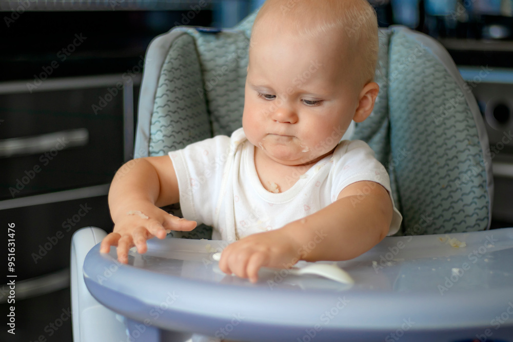 Cute blond caucasian baby child with blue eyes sitting at highchair after eating food. Child is dirty and everything is in rice grains. Baby is serious and surprised Cute blond caucasian baby child with blue eyes sitting at highchair after eating food. Child is dirty and everything is in rice grains. Baby is serious and surprised