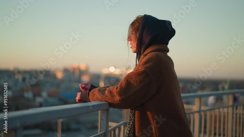 A sorrowful woman walks slowly while holding onto a metal railing, dressed in a brown coat and a hoodie, with a melancholic expression as the cityscape blurs in the background during sunset
