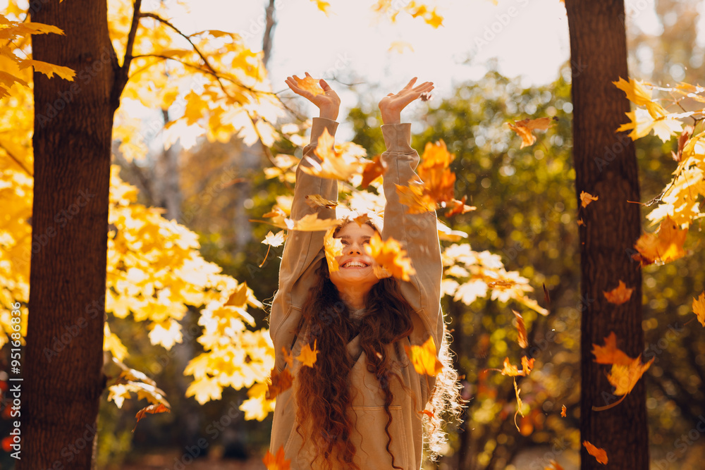 Smiling young woman with hands up throws up the autumn leaves in the forest at sunset