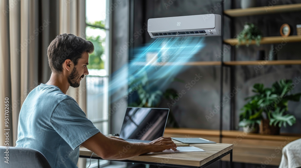 Man working on a laptop in a modern home office, enjoying a cool breeze from an air conditioner Man working on a laptop in a modern home office, enjoying a cool breeze from an air conditioner