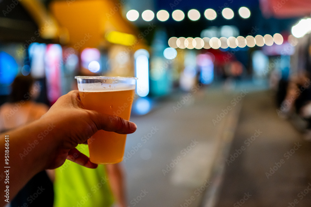 A close-up of a hand holding a plastic cup filled with beer, with a blurred street scene in the background at night. The image captures a casual, urban nightlife atmosphere A close-up of a hand holding a plastic cup filled with beer, with a blurred street scene in the background at night. The image captures a casual, urban nightlife atmosphere
