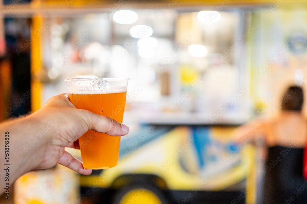 A close-up of a hand holding a glass of beer with a blurred background featuring a food truck. The image captures the essence of enjoying a refreshing drink at an outdoor food festival A close-up of a hand holding a glass of beer with a blurred background featuring a food truck. The image captures the essence of enjoying a refreshing drink at an outdoor food festival