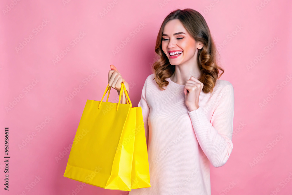 A happy woman in a pink sweater with wavy brown hair holds bright yellow shopping bags. The background is light pink, emphasizing her cheerful mood