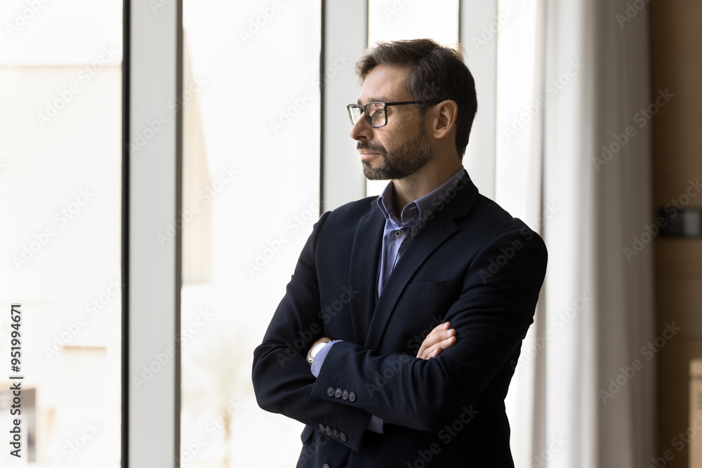 Thoughtful serious company executive man in formal jacket and glasses standing at office window with hands folded, looking away, thinking on real estate investment, future project Thoughtful serious company executive man in formal jacket and glasses standing at office window with hands folded, looking away, thinking on real estate investment, future project