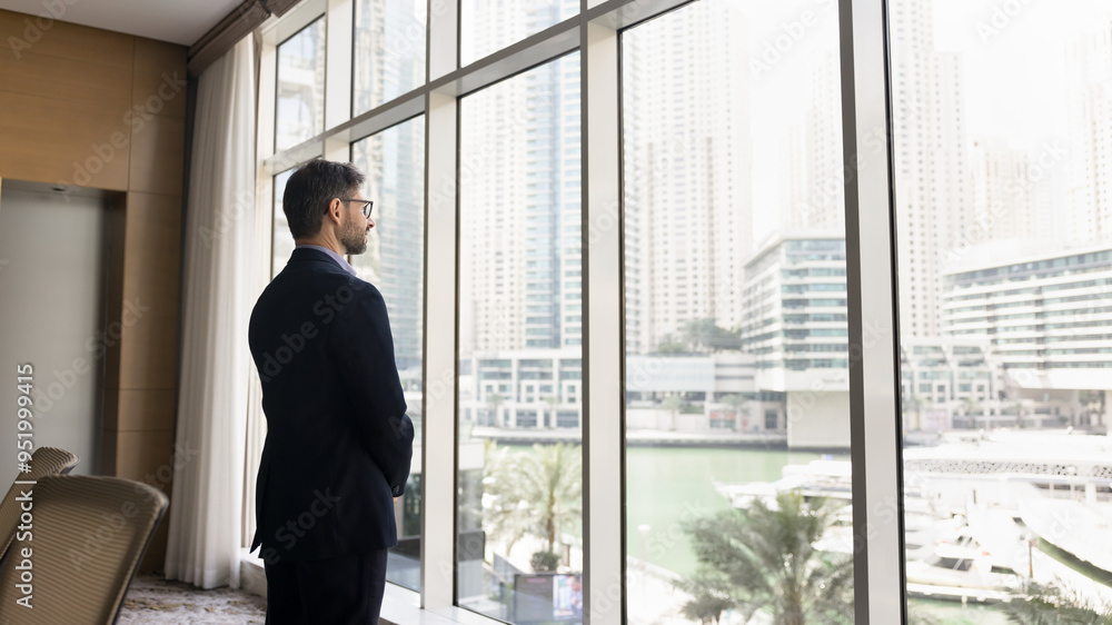 Serious confident business owner man in suit standing at large office window, looking at city view, thinking on real estate investment, property development, professional success Serious confident business owner man in suit standing at large office window, looking at city view, thinking on real estate investment, property development, professional success
