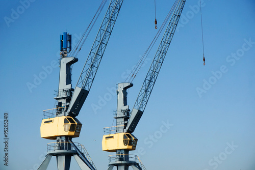 Two large, rusty, metal cranes in a port with hanging chains. Industrial machinery harbor equipment. Clear blue sky in background. Empty copy space industry equipment.