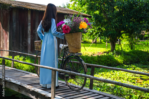 Rear view Vietnamese woman in AO DAI traditional costume standing on wood bridge with flower basket on vintage bicycle in the park