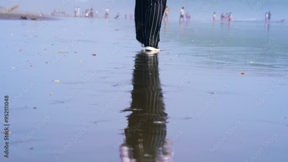 Indian woman wearing trendy shoes and pants walking on a beach with wet ground reflecting her and out of focus crowd in background in morjim in Goa India Indian woman wearing trendy shoes and pants walking on a beach with wet ground reflecting her and out of focus crowd in background in morjim in Goa India
