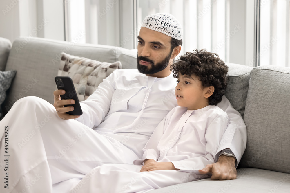 Serious Arabic dad and curious cute little son in white male Muslim dresses using online application on mobile phone, sitting close on home couch, enjoying online and offline communication Serious Arabic dad and curious cute little son in white male Muslim dresses using online application on mobile phone, sitting close on home couch, enjoying online and offline communication