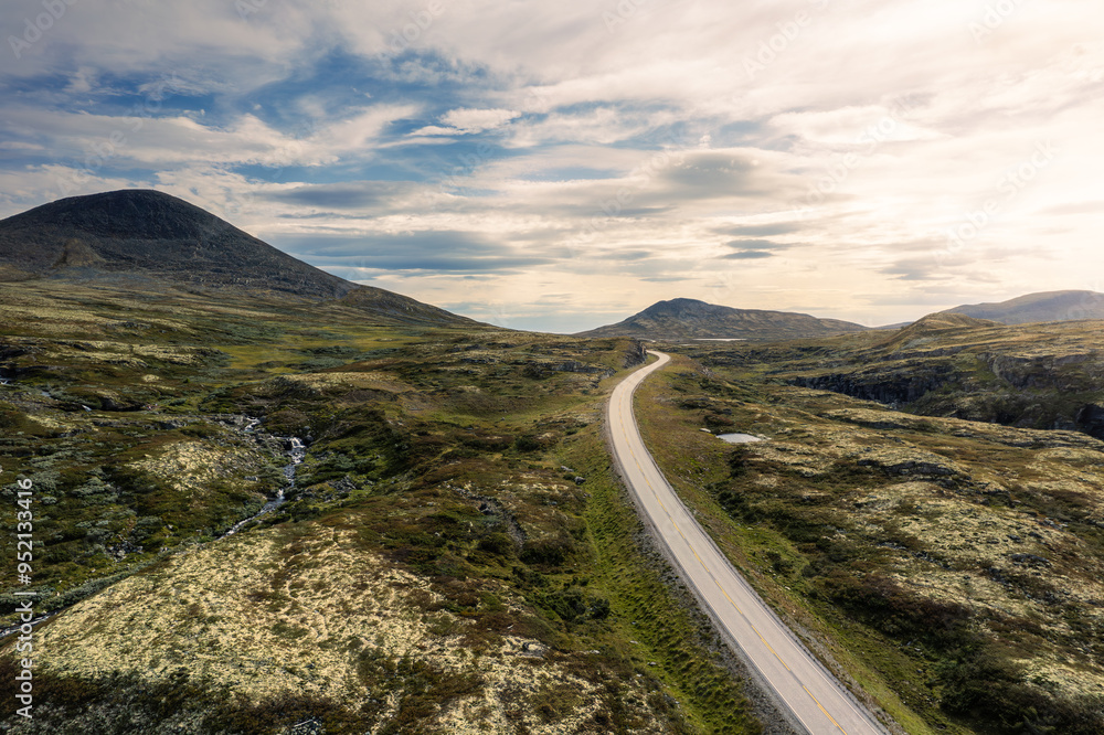 Bright sunlight illuminates the Muen and Nørdre Bølhøgda mountains as a winding road and mountain stream flow through the lush green landscape of Rondane National Park in Norway Bright sunlight illuminates the Muen and Nørdre Bølhøgda mountains as a winding road and mountain stream flow through the lush green landscape of Rondane National Park in Norway