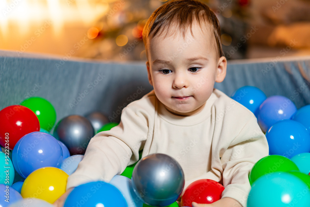 Toddler in a colorful ball pit at home for the holidays. A young child enjoys playing in a vibrant ball pit filled with various colored balls while surrounded by holiday decorations. Toddler in a colorful ball pit at home for the holidays. A young child enjoys playing in a vibrant ball pit filled with various colored balls while surrounded by holiday decorations.