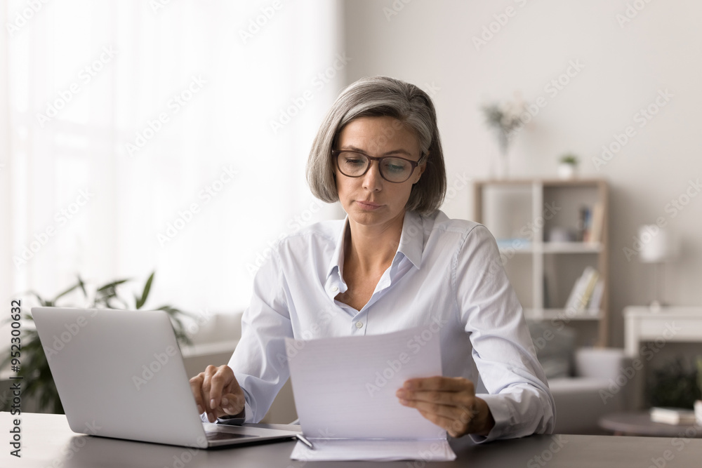 Serious pensive middle-aged woman hold paper document, consider financial report working remotely from home office, work online on laptop, read and analyse contract, reviewing sales data statistics Serious pensive middle-aged woman hold paper document, consider financial report working remotely from home office, work online on laptop, read and analyse contract, reviewing sales data statistics