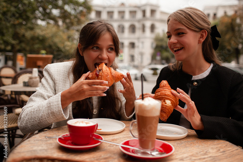 Two beautiful young girls are sitting in a French cafe, eating a croissant and drinking coffee with whipped cream. Teenagers are dressed in business style, black and white jacket in Old Money style
