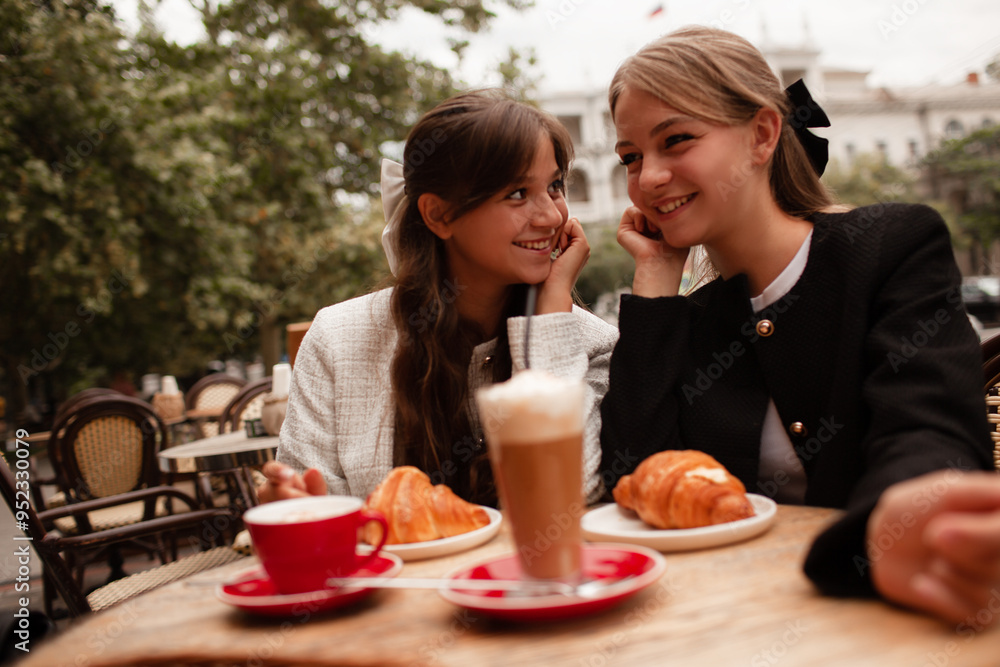 Two beautiful young girls are sitting in a French cafe, eating a croissant and drinking coffee with whipped cream. Teenagers are dressed in business style, black and white jacket in Old Money style Two beautiful young girls are sitting in a French cafe, eating a croissant and drinking coffee with whipped cream. Teenagers are dressed in business style, black and white jacket in Old Money style