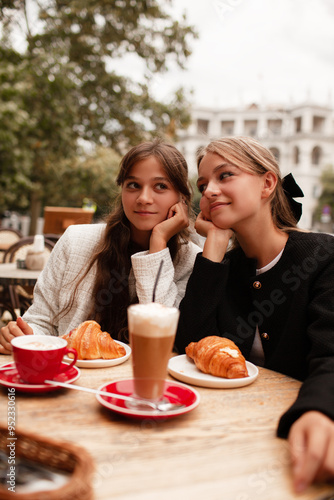 Two beautiful young girls are sitting in a French cafe, eating a croissant and drinking coffee with whipped cream. Teenagers are dressed in business style, black and white jacket in Old Money style