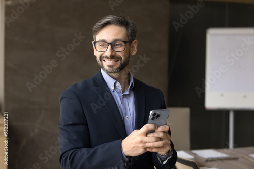 Happy middle aged Hispanic CEO man in glasses holding mobile phone, standing in office board room alone, looking away, smiling, enjoying wireless online communication, thinking on leadership, success