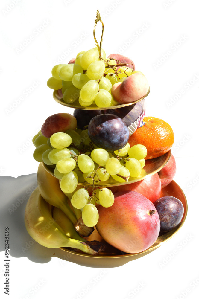 Decadent gold tiered fruit platter isolated on white background, apple, banana, grapes, mandarine orange, fig, mango, plum, detailed, close up. Decadent gold tiered fruit platter isolated on white background, apple, banana, grapes, mandarine orange, fig, mango, plum, detailed, close up.