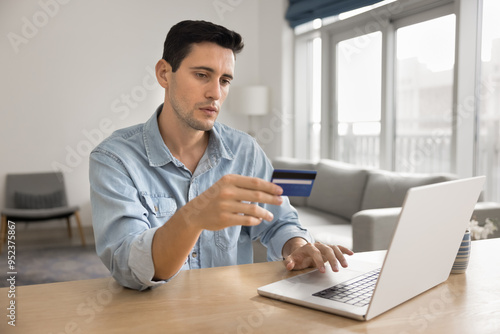 Serious young Hispanic bank client man making money transaction, using blue credit card and online bank financial application, typing on laptop computer, paying for web purchase