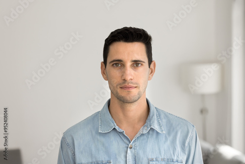 Serious handsome young Latin man posing for front head shot at home with white wall in background, looking at camera. Attractive male Hispanic model guy in casual denim standing indoors for portrait