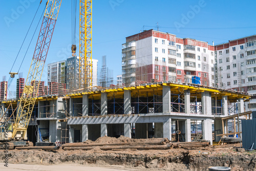 A construction site where tower and crawler cranes perform lifting work. A new house is being built on the site of the old houses. A road is being paved near the house under construction.