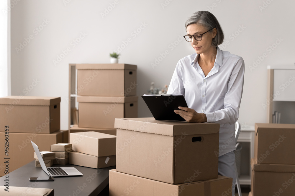 Mature woman, small business owner working in stockroom filled with cardboard boxes, using clipboard to check details related to packages or shipment details, involved in logistics, inventory workflow Mature woman, small business owner working in stockroom filled with cardboard boxes, using clipboard to check details related to packages or shipment details, involved in logistics, inventory workflow