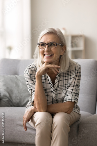 Vertical portrait of attractive senior woman resting on couch, wear glasses, look into distance, deep in pleasant thoughts, contemplate, daydreaming spends weekend sit on sofa. Retirement and comfort