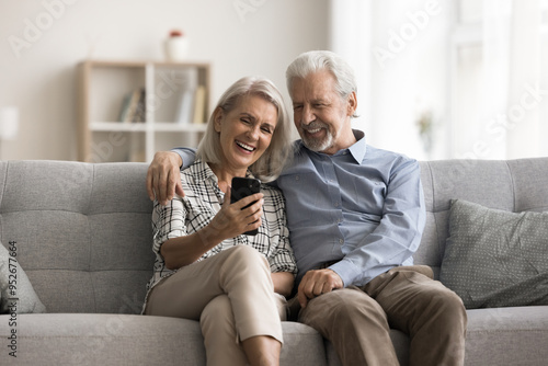 Happy aged wife and husband sit on sofa look at mobile phone screen, laughing, watching funny videos in social media, spend carefree pastime on internet using gadget. Olde gen and modern tech usage photo