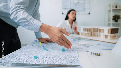 Skilled caucasian interior designer working with house model while discussing with asian architect engineer about house plan on meeting table with blueprint and house model scatter around. Immaculate. photo