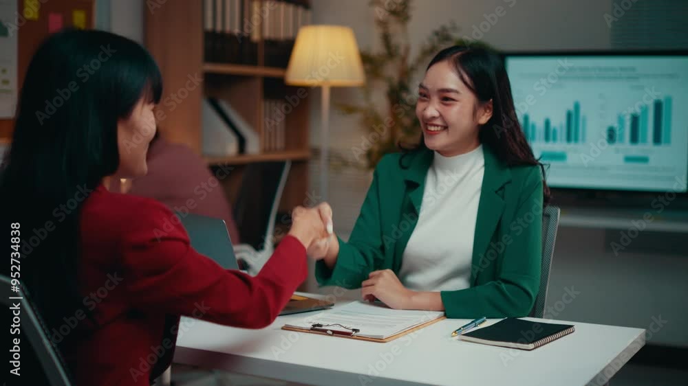 Two asian businesswomen are meeting in an office. One is wearing a green blazer and the other a red one. They are shaking hands above a desk Two asian businesswomen are meeting in an office. One is wearing a green blazer and the other a red one. They are shaking hands above a desk
