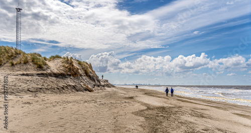 A sandy beach stretches along the Zandmotor shoreline near Kijkduin, with a couple walking near the water's edge under a partly cloudy sky. Dunes and a tall antenna tower rise on the left.