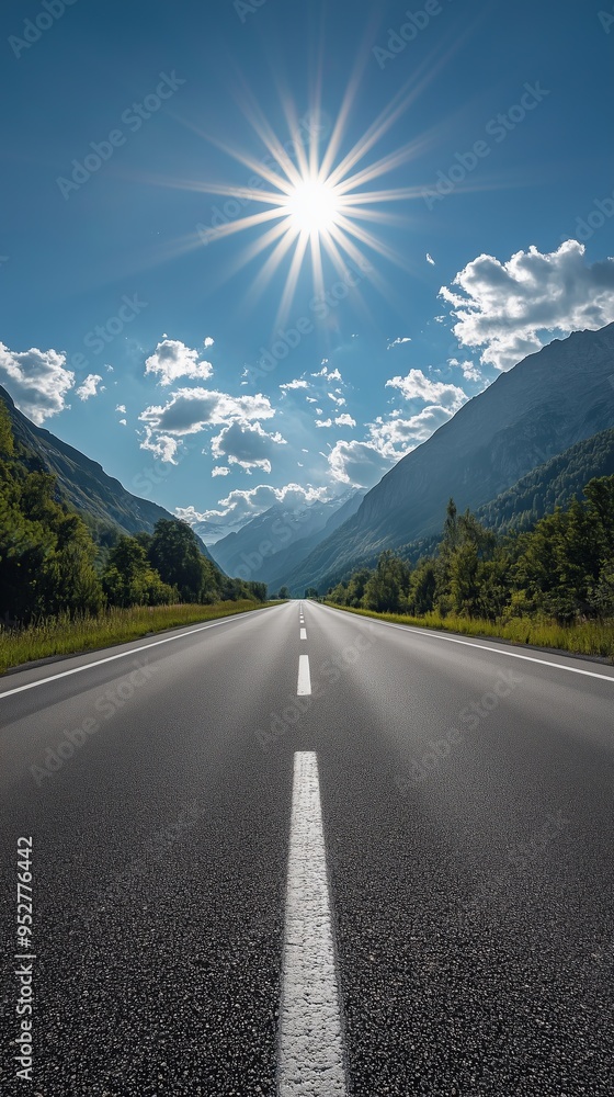 Fototapeta premium Straight asphalt road leading through mountainous terrain under a bright sun and clear blue sky with scattered clouds.