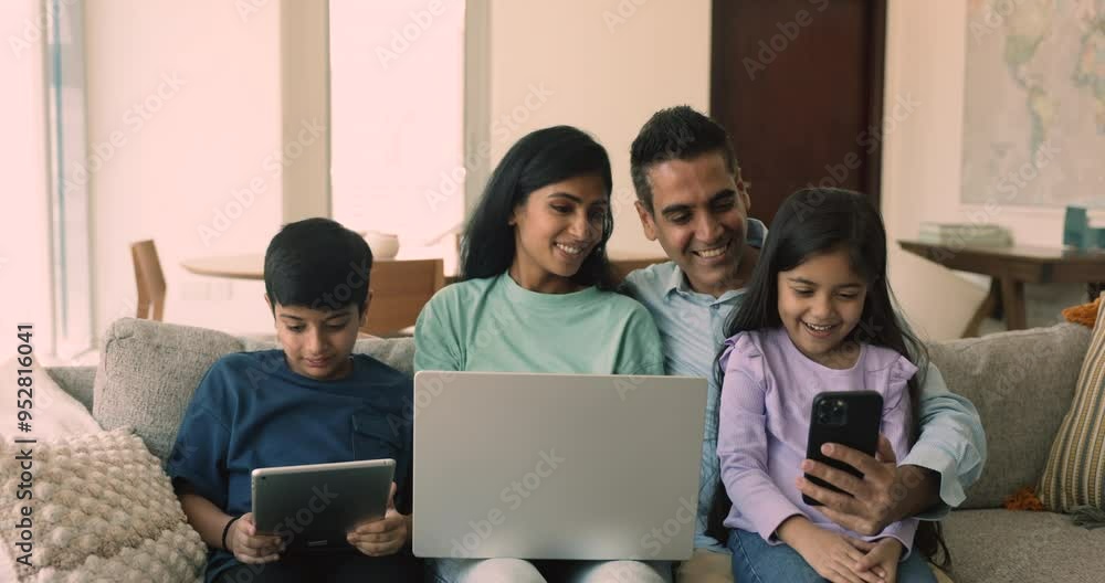 Happy young Indian parents and two sibling kids holding digital gadgets, sitting close together on home couch, using Internet application, media service on smartphone, tablet, laptop, talking, smiling Happy young Indian parents and two sibling kids holding digital gadgets, sitting close together on home couch, using Internet application, media service on smartphone, tablet, laptop, talking, smiling