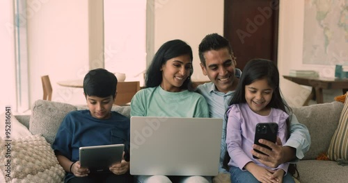 Happy young Indian parents and two sibling kids holding digital gadgets, sitting close together on home couch, using Internet application, media service on smartphone, tablet, laptop, talking, smiling photo