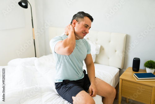 Hispanic man sitting on bed at home, experiencing neck stiffness and touching his painful neck