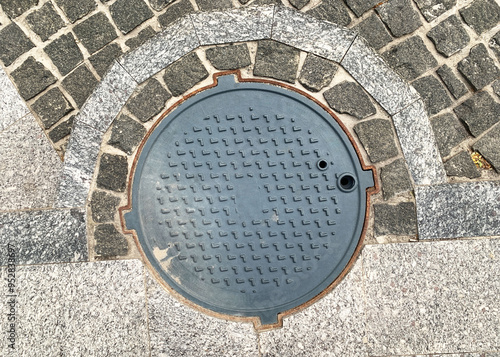 Sewer black round cast iron manhole cover embedded in a cobblestone pavement, showcasing a geometric pattern and weathered texture. Top view. Close up