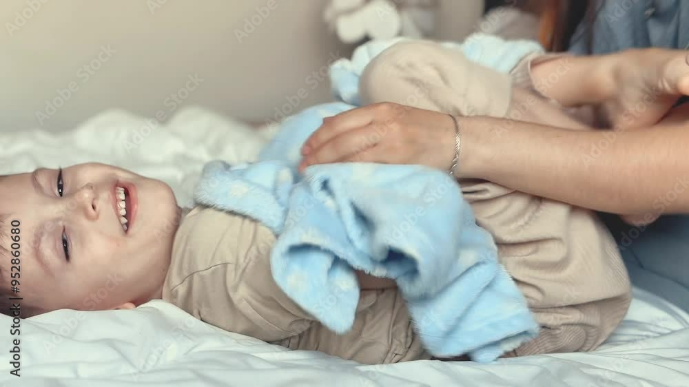 Mom tickles the feet of her little son, wrapped in a blue blanket. Mom and child have fun together in a cozy bedroom at home. Happy children with mother. Bright photos of the baby's childhood. Mom tickles the feet of her little son, wrapped in a blue blanket. Mom and child have fun together in a cozy bedroom at home. Happy children with mother. Bright photos of the baby's childhood.