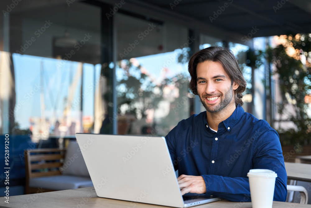 Fototapeta premium Happy young man user entrepreneur online business owner using computer sitting outdoors in coffee shop. Smiling guy student or professional looking at camera in cafe hybrid working on laptop, portrait
