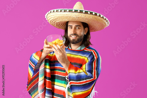 Handsome young Mexican man in sombrero, poncho and with nachos on magenta background