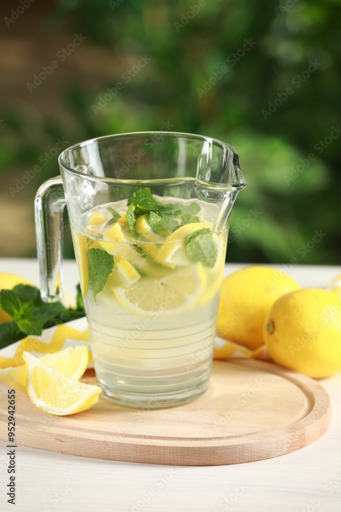 Refreshing lemonade with mint in jug and fruits on light table against blurred green background
