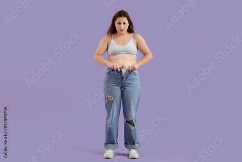 Overweight young woman in tight jeans on purple background. Weight gain concept photo