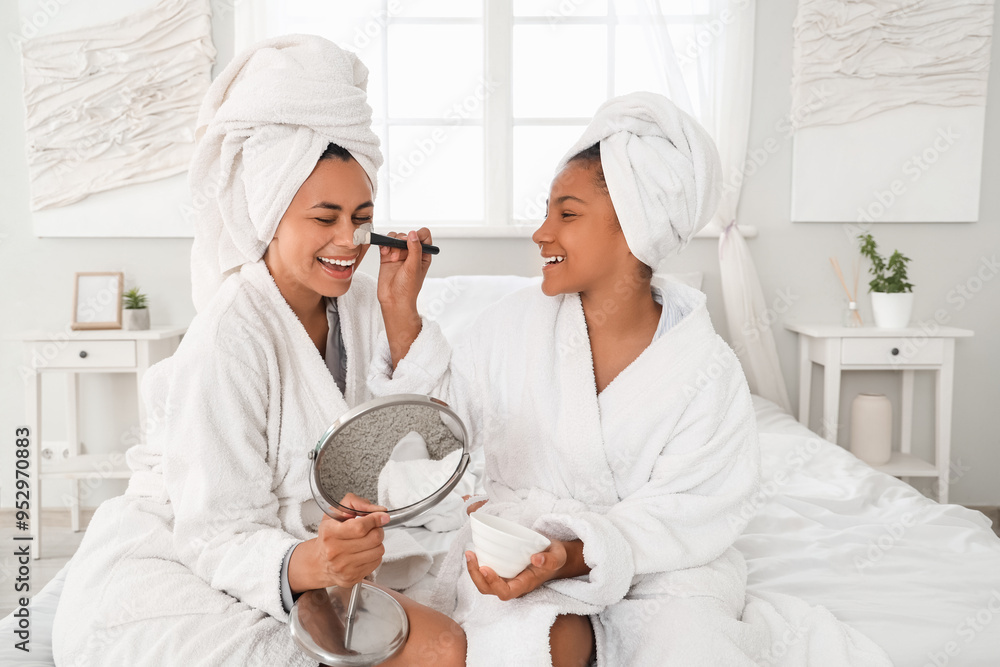 Fototapeta premium Happy young African-American mother and her daughter in bathrobes with mirror applying facial mask at home